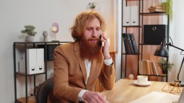 Genç işadamı ofis masasında oturuyor cep telefonuyla müşteriye cevap veriyor, akıllı telefon kullanıyor. Sohbet. Kafkasyalı kızıl saçlı, uzaktan kumandalı bir işte çalışıyor..