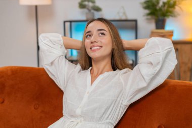 Dertsiz, genç, beyaz bir kadın evdeki oturma odasında elleri başının arkasında, koltukta rahatça oturuyor. Kanepede dinlenen gülümseyen kız daire odasına bakınmayı düşünüyor. Yaşam biçimi.