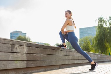 Koşu egzersizlerinden önce bacaklarının esnediği atletik genç bir kadının yan görüntüsü. Beyaz kadın koşucu kız güneşli bir günde şehir parkında hamleler yapıyor. Sağlıklı yaşam tarzı kavramı.