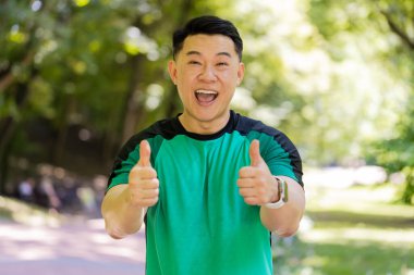 Gibi. Portre atletik formda gülümseyen koşucu eğitimi kameraya bakıp maraton başarısına seviniyor. Parkta egzersiz kardiyolojisi. Japon genç sporcu bulanık ağaçlara karşı baş parmağını kaldırıyor