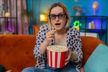 Kanepede oturup patlamış mısır yiyen ve ilginç TV dizileri, spor oyunları, filmler, evdeki sosyal medya içerikleri izleyen heyecanlı genç bir kadın. Üç boyutlu gözlüklü kız ev eğlencesi yapıyor.