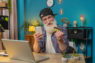 Merkez ofisteki online alışveriş ödemeleri için banka kartı ve dizüstü bilgisayar kullanan Kafkasyalı mutlu yaşlı bir adam. İş anlaşması. Gülümseyen şık büyükbaba satın alımları netbook E-bankacılık uygulama hizmeti ile.