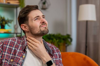 Ekose gömlekli, üzgün, beyaz bir adam boğaz ağrısı için el ele tutuşuyor. Ağrılı boyun ve kaşları çatık, tiroid bozuklukları boğaz ağrısı, bademcik iltihabı. Sınıftaki kanepede oturan genç adam.