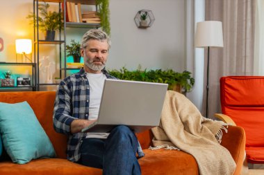 Katı sakallı, olgun bir adam dizüstü bilgisayarda yazı yazıyor. İnternette geziniyor. Kanepede tek başına oturan uygulamaları kullanıyor. Orta yaşlı, yaşlı, beyaz bir adam bilgisayarda çalışıyor. Oturma odasında ekrana bakıyor.