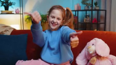 Gibi. Mutlu genç kız kameraya onaylayıcı bir şekilde bakıyor olumlu bir işaret veriyor, olumlu bir geri bildirim. Genç bir kız, akşam evde, oturma odasında, kanepede oturuyor.