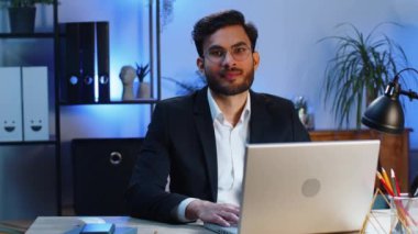 Modern ofis çalışma masasındaki mutlu, sakallı Hintli işadamının portresi başını kameraya çevir. Resmi takım elbiseli bir erkek Hindu. Yakışıklı Uzaktan kumandalı adam dizüstü bilgisayarla çalışıyor