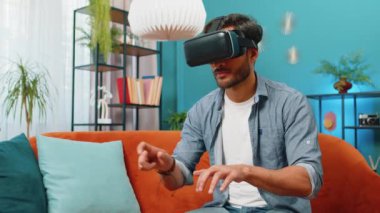 Hintli adam sanal gerçeklik teknolojisi VR uygulama başlığı kullanarak sanal klavyeye yazı yazarak, evdeki dairede 3D video oyunu simülasyonu yaparak. Kanepede oturan gözlüklü adam.