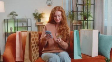 Alışveriş meraklısı kızıl saçlı mutlu bir kadın alışveriş torbalarıyla evde oturuyor online sipariş veriyor, kredi kartı ile ödeme yapıyor. Evdeki koltukta oturan heyecanlı kadın...