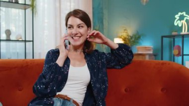 Gülümseyen beyaz bir kadın evdeki kanepede oturan arkadaşlarıyla telefonda konuşuyor. Gündelik kıyafetlerle mutlu bir kız modern apartman dairesinde dedikodu yapıyor..