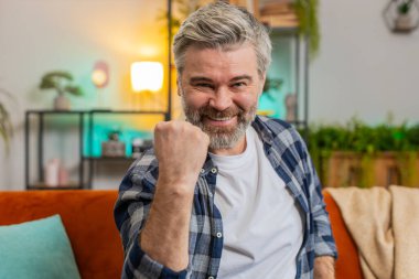 Mutlu Kafkas kıdemli adam TV izlerken bağırıyor, başarıyı kutluyor, kazanıyor, gol başarısı, iyi haber, piyango şansı, oturma odasında koltukta oturan zafer. Evde yumruklarını sıkan olgun bir ihtiyar.