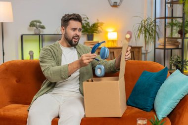 Mutsuz genç beyaz adam kutuları açıyor, kablosuz kulaklık siparişinden memnun değil, mobil uygulamalara olumsuz geri bildirim yazıyor, internet alışverişi kavramı.