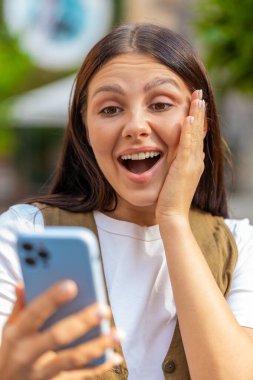 Aman Tanrım. Vay canına. Heyecanlı mutlu, neşeli Avrupalı genç kadın akıllı telefon daktilo kullanıyor kutlama yapıyor açık havada iyi haber kazan. Şehir caddesinde duran yetişkin kız. Kasaba yaşam tarzları