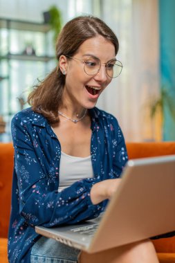 Gözlüklü, dizüstü bilgisayar kullanan, iyi haber mesajı alan mutlu genç bir kadın aniden kazanılan piyango ikramiyesini kutlamak için ağzını açtı. Evden online alışveriş yapıyor..