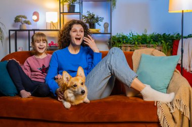 Şaşırmış anne kız ve kız çocuğu Corgi Dog 'un yanında oturuyor TV seyrediyor evde gülüyor. Mutlu bir aile, en sevdiği TV dizisini izleyip hafta sonu boyunca koltukta oturur..
