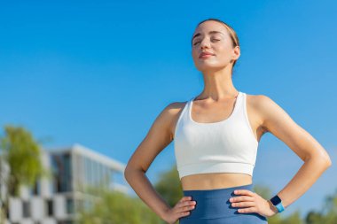 Şehir parkında sürat koşusundan sonra genç beyaz kadın koşucu nefes darlığı çekiyor. Elleri kalçalarında gezen ve yaz günü derin derin nefes alan formda bir bayan sporcu. Sağlıklı yaşam tarzı.
