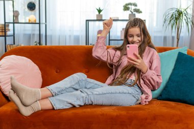 Mobil akıllı telefon yazma oyunu oynayan mutlu bir kız büyük bir sürpriz buldu, iyi haberler, online maç kazancını kutladı. Oturma odasında el çırpan genç kız çocuk kanepede oturuyor..