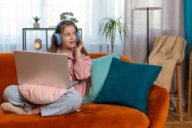 Laptop kullanan, kulaklıklı beyaz ergen bir kız müzik dinliyor ya da ders dinliyor, uzaktan öğreniyor. Online eğitim. Gülümseyen küçük çocuk dinleniyor, evinde dinleniyor kanepede oturuyor..