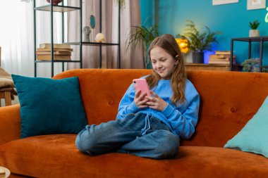 Neşeli genç kız, internet üzerinden akıllı telefon sosyal medya uygulamalarında rahatlama filmi izlerken mesajlarını paylaşıyor. Oturma odasında cep telefonu kullanan beyaz bir kız çocuk kanepede oturuyor..