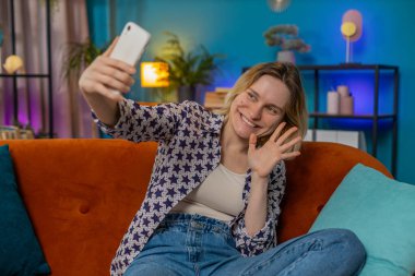 Kafkasyalı mutlu bir kadın akıllı telefondan video görüşmesi yapıyor ve el sallayarak selam veriyor. İletişim, toplantı. Gülümseyen kız blogcu cep telefonuyla konuşuyor. Evdeki turuncu koltukta oturuyor.