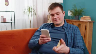 Para transfer ederken kredi kartı ve akıllı telefon kullanan beyaz bir adam, kapalı bir evde çevrimiçi alışveriş nakitsiz yemek siparişi veriyor. Mutlu genç adam kanepede oturuyor..