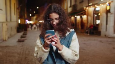 Gülümseyen yorgun genç kız akıllı telefon kullanıyor internette mesajlar yazıyor, işi bitiriyor, kameraya bakıyor. Geceleri şehir caddesinde dikilen bir kadın. Şehir dışında..
