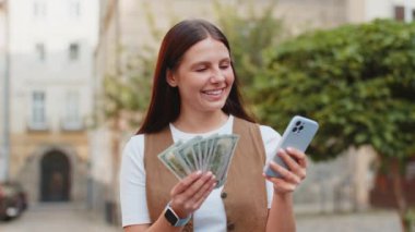 Mutlu genç bir kadın nakit para sayıyor, akıllı telefon hesap makinesi uygulaması kullanıyor, planlanmış tatil hediyeleri için gelirden tatmin oluyor. Şehir merkezinde güneşli bir sokakta duran güzel bir kız. Şehir yaşam tarzları açık havada
