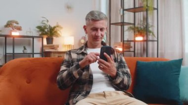 Evdeki oturma odasında cep telefonu tebessümleri kullanarak kanepede oturan beyaz bir genç. Gri saçlı mutlu adam mesajlaşırken akıllı telefon sosyal medya uygulamaları içeriğini paylaşıyor.
