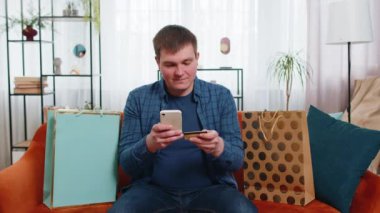Apartmanda alışveriş torbalarıyla oturan, online sipariş veren, kredi kartı ile ödeme yapan mutlu tüketici. Genç adam evdeki kanepede sipariş alıyor. İnternette indirimli satış.