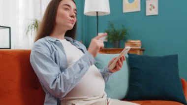 Akıllı telefon mesajlarıyla gülümseyen hamile bir kadın sosyal medya sohbetleriyle evdeki oturma odasındaki kanepede oturuyor. Mutlu beyaz kadın gelecekteki anne bebek için internetten alışveriş yapıyor.