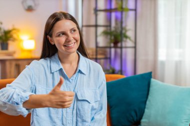 Heyecanlı gülümseyen beyaz kadın portresi onaylayıcı bir şekilde kameraya bakıyor işaret, iyi haber, olumlu geri bildirim gibi. Evdeki oturma odasında kanepede oturan mutlu genç kız..