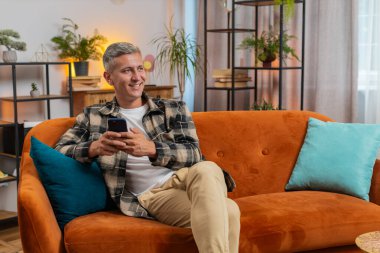 Evdeki oturma odasında cep telefonu tebessümleri kullanarak kanepede oturan beyaz bir genç. Gri saçlı mutlu adam mesajlaşırken akıllı telefon sosyal medya uygulamaları içeriğini paylaşıyor.