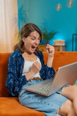 Laptop kullanan Kafkasyalı genç kadın, iyi haberler aldı. Aniden kazanılan zaferle şok oldu. Dairede piyango ikramiyesini kutladı. Neşeli kız evdeki salonda kanepede çığlık atıp dans ediyor.