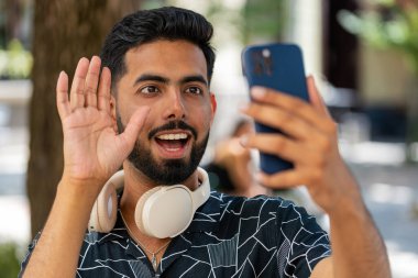 Akıllı telefon kamerasına gülümseyen neşeli genç turist el sallayarak merhaba, merhaba, merhaba, güle güle, misafirperver bir yüz ifadesiyle hoş geldiniz diyor. Şehir sokağındaki Hintli adam..