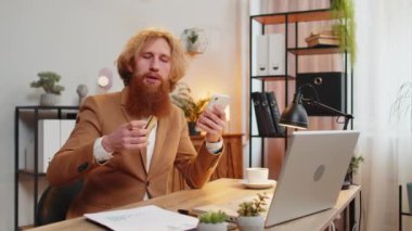 Merkez ofisteki online alışveriş ödemeleri için banka kartı ve akıllı telefon kullanan mutlu bir beyaz adam. İş anlaşması. Gülümseyen kızıl saçlı, cep telefonu E-bankacılık uygulama servisiyle alım yapıyor..