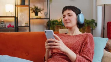 Kablosuz kulaklıklı mutlu beyaz kız akıllı telefondan en sevilen enerjik dans müziğini dinliyor. Gülümseyen genç kadın hafta sonu boyunca evdeki oturma odasındaki kanepede dinleniyor..