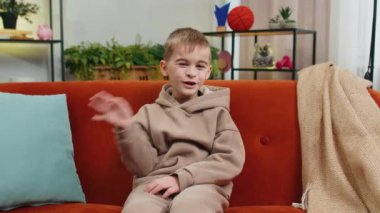 Kameraya gülümseyen ve el sallayan genç çocuk merhaba, merhaba, merhaba ya da güle güle diyerek misafirperver bir yüz ifadesiyle karşıladı. Oturma odasındaki beyaz çocuk kanepede oturuyor.