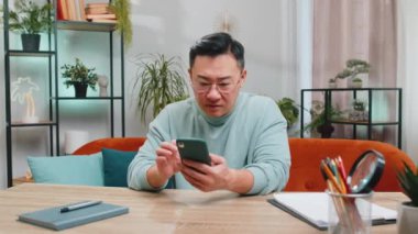 Hüzünlü Çinli adam akıllı telefon daktilo kullanıyor, beklenmedik piyango sonuçlarını kaybediyor, kötü haber, servet kaybı, son teslim tarihi, virüs. Olgun Asyalı çocuk kanepede oturuyor.