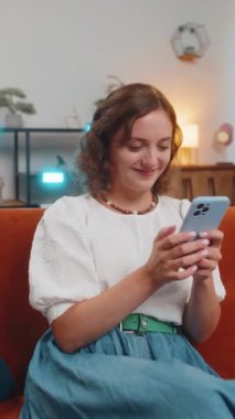 Oturma odasında gülümseyen cep telefonuyla kanepede oturan genç kadının portresi. İnternet üzerinden akıllı telefon sosyal medya uygulamaları üzerinden mesajlaşan bir kız. Dikey.