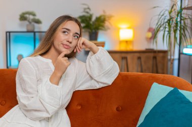 Beyaz elbiseli, düşünceli, beyaz tenli, çenesine dokunarak hafta sonu için nereye gideceğini düşünen, evdeki oturma odasındaki kanepede oturan yeni fikirler üreten genç, çekici beyaz bir kadın. Yaşam biçimi