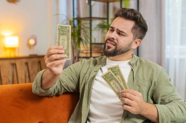 Zavallı Kafkasyalı genç adam yetersiz para için üzülüyor, dolar banknotlarını sayıyor. Finansal kriz. İflas. Yoksulluk ve yoksulluk. Evdeki kanepede oturan üzgün yetişkin adam..