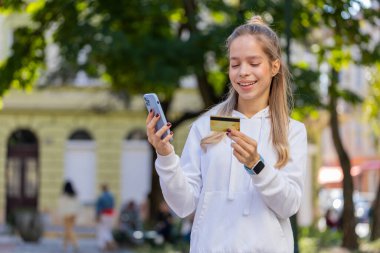 Para transfer ederken kredi kartı akıllı telefonu kullanan gülümseyen bir kadın, online alışveriş, yemek siparişi, açık havada otel rezervasyonu. Şehir caddesinde duran genç kız..