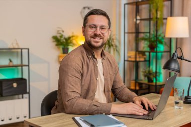 Genç iş adamı dizüstü bilgisayarla oturma odasında kameraya gülümsüyor. Happy Kafkasyalı adam serbest çalışıyor. Ev masasında uzaktan kumandalı çalışıyor. Bilgisayarda E-öğrenme, internette gezinme.