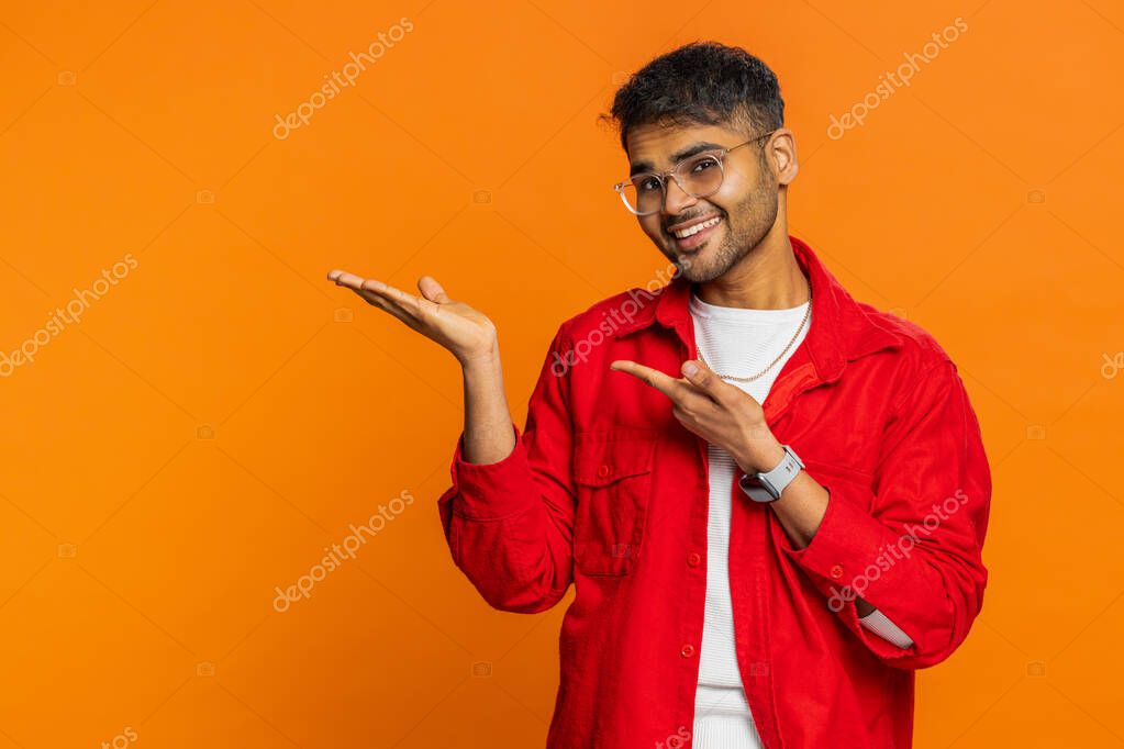Indian man showing thumbs up and pointing left empty place, advertising area for commercial text copy space for goods promotion advertisement. Arabian guy isolated on orange background. Lifestyles
