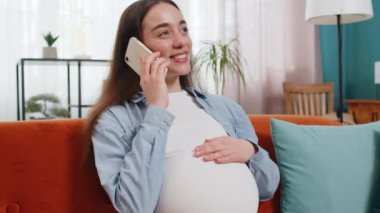 Oturma odasındaki koltukta oturmuş akıllı telefondan konuşan hamile bir kadın. Evinde cep telefonu tutan mutlu bir beyaz kadın. Doğum öncesi bakım ve hamilelik konsepti.