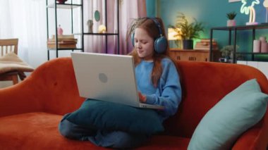 Laptop kullanan, kulaklıklı beyaz ergen bir kız müzik dinliyor ya da ders dinliyor, uzaktan öğreniyor. Online eğitim. Gülümseyen küçük bir çocuk, dinlenen bir çocuk, iş yerinde kanepede oturuyor..