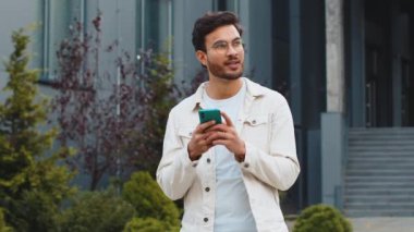 Genç gülümseyen Hintli adam akıllı telefon kullanıyor kısa mesajlar yazıyor, e-posta okuyor, dışarıda haberleri inceliyor. Şehir merkezindeki caddede duran Arap Hindu adam mobil sosyal medya uygulaması kullanıyor. İş adamları