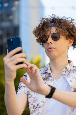 Mutlu, gülümseyen esmer adam internet sosyal medya uygulamalarını karıştırarak açık havada sohbet ederek akıllı telefon yazıları yazıyor. Şehir sokağında kıvırcık saçlı bir turist. Yaşam Tarzları.