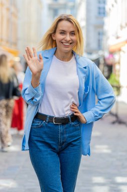 Neşeli genç bayan turist kameraya gülümsüyor, el sallıyor, el sallıyor merhaba, selam, selam, hoşçakal, misafirperver bir yüz ifadesiyle hoş geldiniz. Şehir merkezindeki kadın.