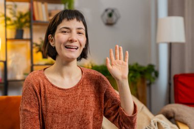 Kameraya gülümseyen ve el sallayan beyaz bir kadının portresi merhaba diyor, evdeki kanepede otururken konuşuyor. Mutlu kız evde vlog kaydediyor.