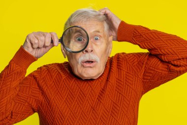 Araştırmacı bilimadamı kıdemli yaşlı adam elinde büyüteçle kameraya bakıyor, aranıyor, analiz ediyor. Sarı duvar arkasında olgun büyükbaba